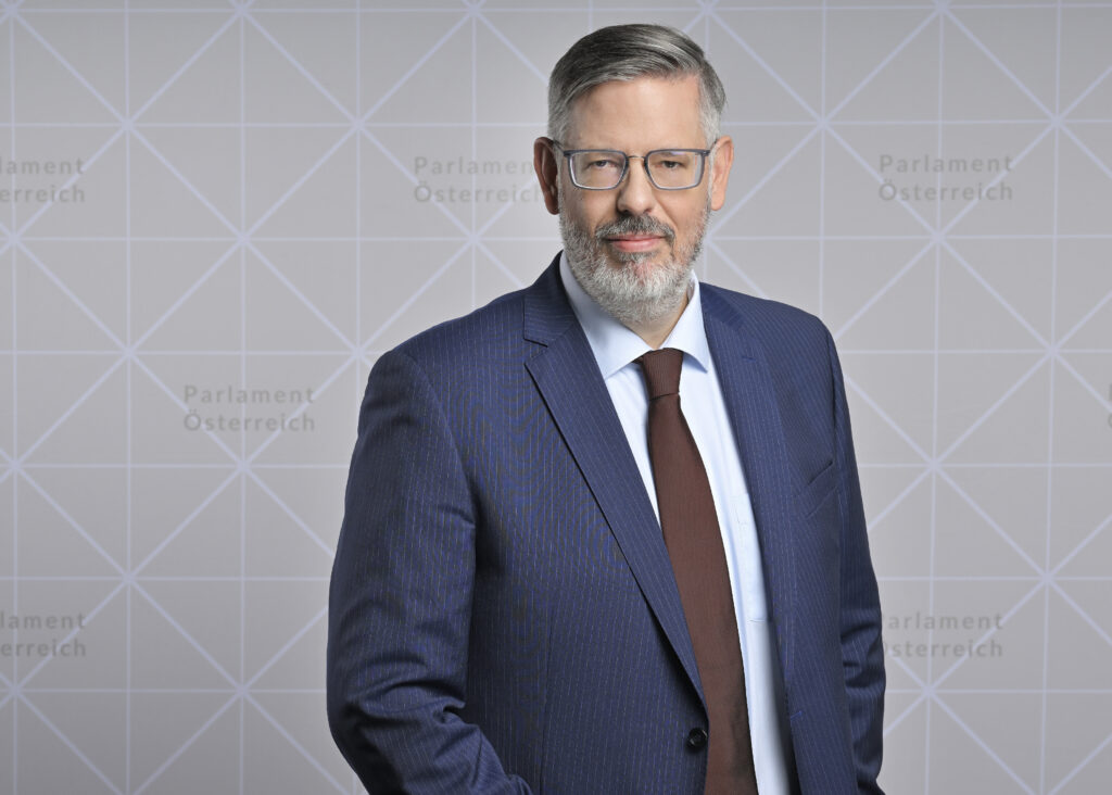 Ralph Schallmeiner, Nationalratsabgeordneter, steht vor einem Hintergrund mit dem Text "Parlament Österreich". Er trägt einen blauen Nadelstreifenanzug, ein hellblaues Hemd und eine dunkelrote Krawatte. Schallmeiner hat graues Haar, eine Brille und einen gepflegten Bart. Er lächelt leicht und blickt direkt in die Kamera.