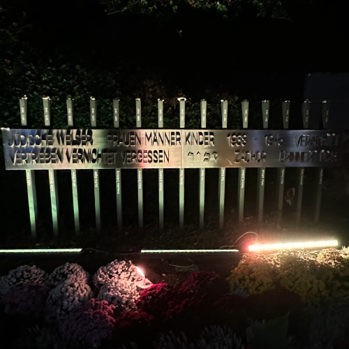 Ein Denkmal bei Nacht, das aus mehreren beleuchteten vertikalen Metallsäulen besteht. In der Mitte befindet sich eine Metallplatte mit deutscher Inschrift “FRAUEN MÄNNER KINDER” und Jahreszahlen, die an die Opfer des Holocaust erinnern. Das Denkmal wird von unten durch rötliches Licht beleuchtet, was eine eindringliche und würdevolle Atmosphäre schafft. Im Vordergrund sind Steine oder Kiesel zu sehen, die im Licht schimmern.