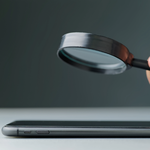Eine Lupe wird über ein Smartphone gehalten, das auf einer dunklen Oberfläche liegt. Die Szene symbolisiert eine genaue Untersuchung oder Analyse des Geräts.