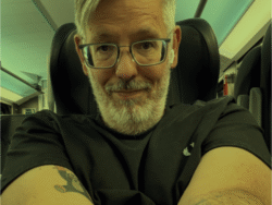 Ein Mann mittleren Alters mit grauem Haar und Bart sitzt in einem modernen Zugabteil. Er trägt eine Brille, ein schwarzes T-Shirt und hat sichtbare Tattoos auf beiden Unterarmen. Der Mann lächelt leicht in die Kamera und macht ein Selfie, während er auf einem bequemen Sitzplatz sitzt. Die Innenbeleuchtung des Zuges ist dezent, und im Hintergrund sind weitere Sitzreihen zu erkennen.