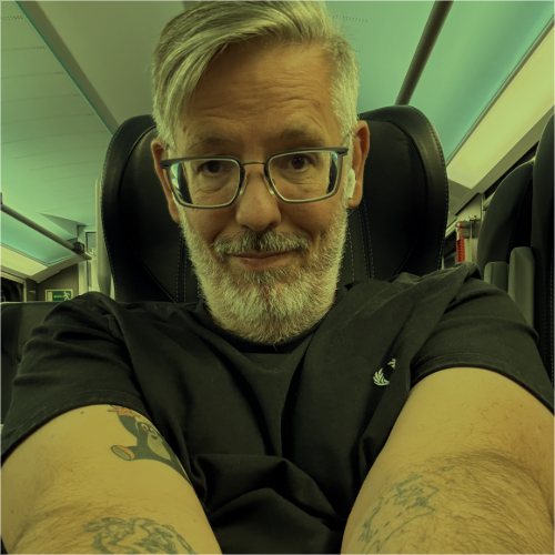 Ein Mann mittleren Alters mit grauem Haar und Bart sitzt in einem modernen Zugabteil. Er trägt eine Brille, ein schwarzes T-Shirt und hat sichtbare Tattoos auf beiden Unterarmen. Der Mann lächelt leicht in die Kamera und macht ein Selfie, während er auf einem bequemen Sitzplatz sitzt. Die Innenbeleuchtung des Zuges ist dezent, und im Hintergrund sind weitere Sitzreihen zu erkennen.