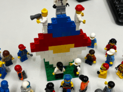 Auf einem weißen Tisch steht eine große, farbenfrohe Lego-Konstruktion in Form eines Pilzes, umgeben von zahlreichen Lego-Männchen. Einige Figuren tragen Berufs- oder Schutzkleidung, eine hält ein Megafon, andere stehen auf der Pilzspitze. Rund um die Konstruktion versammeln sich viele unterschiedliche Lego-Figuren, die eine vielseitige Gruppe darstellen. Im Hintergrund sind Teile einer Tastatur und ein schwarzes Gerät zu sehen.