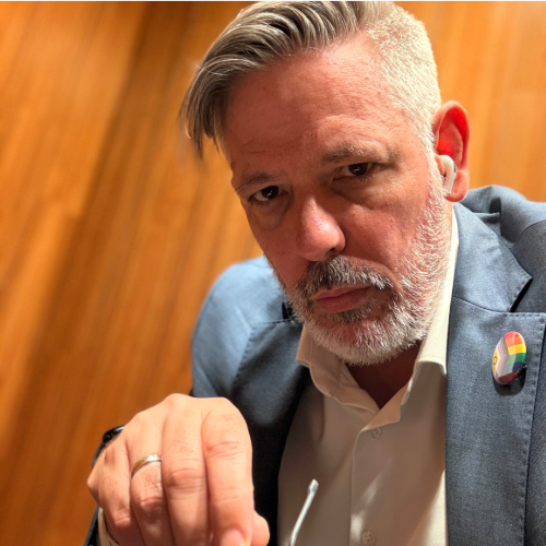 Ein Mann mit grauem Haar und Bart sitzt in einem blauen Anzug und weißem Hemd vor einer braunen Holzwand. Er blickt ernst in die Kamera und trägt eine Anstecknadel mit Regenbogen-Muster am Revers.