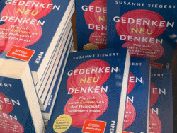 Mehrere Stapel des Buches „Gedenken neu denken – Wie sich unser Erinnern an den Holocaust verändern muss“ von Susanne Siegert liegen auf einem Tisch. Die Bücher haben ein blaues Cover mit einer roten, spiralförmigen Grafik in der Mitte und tragen den Hinweis „SPIEGEL Bestseller“.