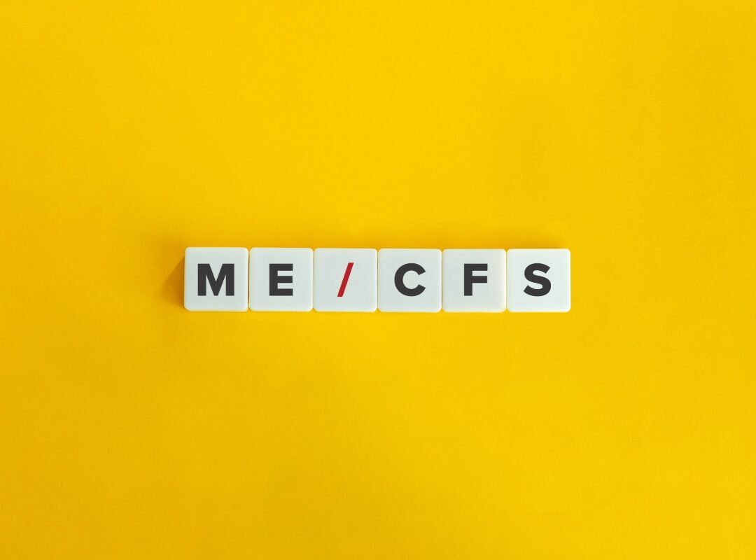 Auf einem gelben Hintergrund liegen sechs weiße Buchstabenwürfel in einer Reihe. Darauf steht „ME/CFS“, wobei der Schrägstrich rot dargestellt ist, während die übrigen Buchstaben schwarz sind. Das Bild ist minimalistisch gestaltet und stellt die Abkürzung für die Erkrankung Myalgische Enzephalomyelitis/Chronisches Fatigue-Syndrom dar.