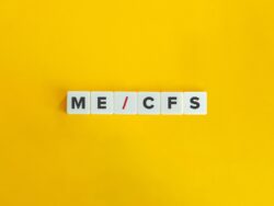 Auf einem gelben Hintergrund liegen sechs weiße Buchstabenwürfel in einer Reihe. Darauf steht „ME/CFS“, wobei der Schrägstrich rot dargestellt ist, während die übrigen Buchstaben schwarz sind. Das Bild ist minimalistisch gestaltet und stellt die Abkürzung für die Erkrankung Myalgische Enzephalomyelitis/Chronisches Fatigue-Syndrom dar.