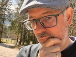 Nahaufnahme eines nachdenklich wirkenden Mannes mit grauem Vollbart, Brille und grauer Schiebermütze, der die Hand ans Kinn legt. Er trägt einen dunklen Pullover. Im Hintergrund sind Bäume und ein sonniger Naturweg zu sehen.