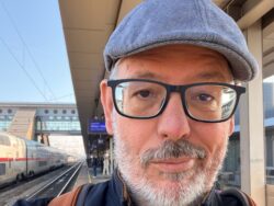 Porträt-Selfie einer Person mit grauer Schiebermütze, schwarzer Brille, Bart und dunkler Jacke auf einem Bahnsteig. Im Hintergrund stehen ein Zug, Gleise und das Bahnhofsgebäude von Wels bei sonnigem Wetter.
