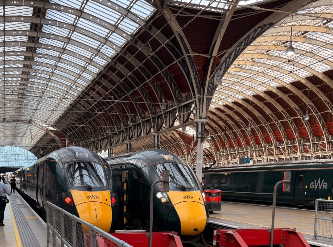 Zwei grün-gelbe Züge stehen nebeneinander unter der imposanten gläsernen Hallenkonstruktion des Londoner Bahnhofs Paddington. Im Hintergrund ist ein weiterer Zug von Great Western Railway zu sehen.