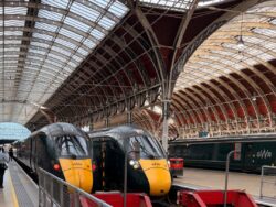 Zwei grün-gelbe Züge stehen nebeneinander unter der imposanten gläsernen Hallenkonstruktion des Londoner Bahnhofs Paddington. Im Hintergrund ist ein weiterer Zug von Great Western Railway zu sehen.